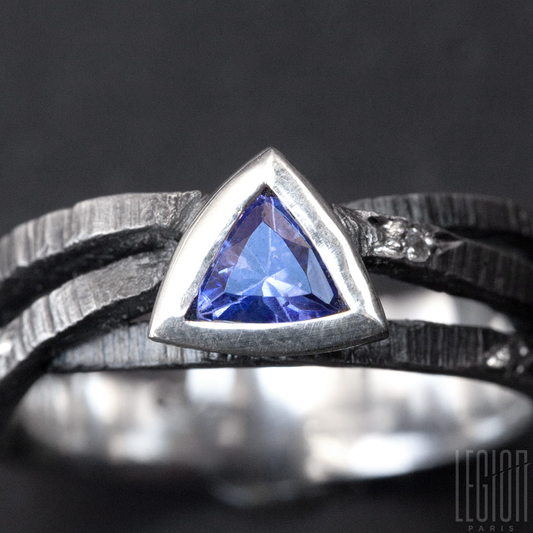 gros plan sur une une bague en argent noir texturé composée de 3 brindilles se croisant sur le dessus et d'une tanzanite bleue de forme triangulaire sertie en clos avec sur le corps de bagues des diamants gris parsemés