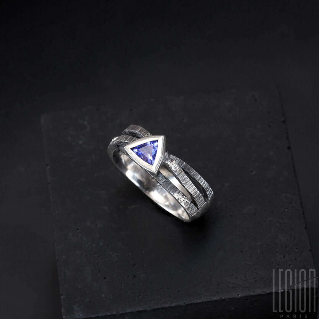une bague en argent noir texturé composée de 3 brindilles se croisant sur le dessus et d'une tanzanite bleue de forme triangulaire sertie en clos avec sur le corps de bagues des diamants gris parsemés