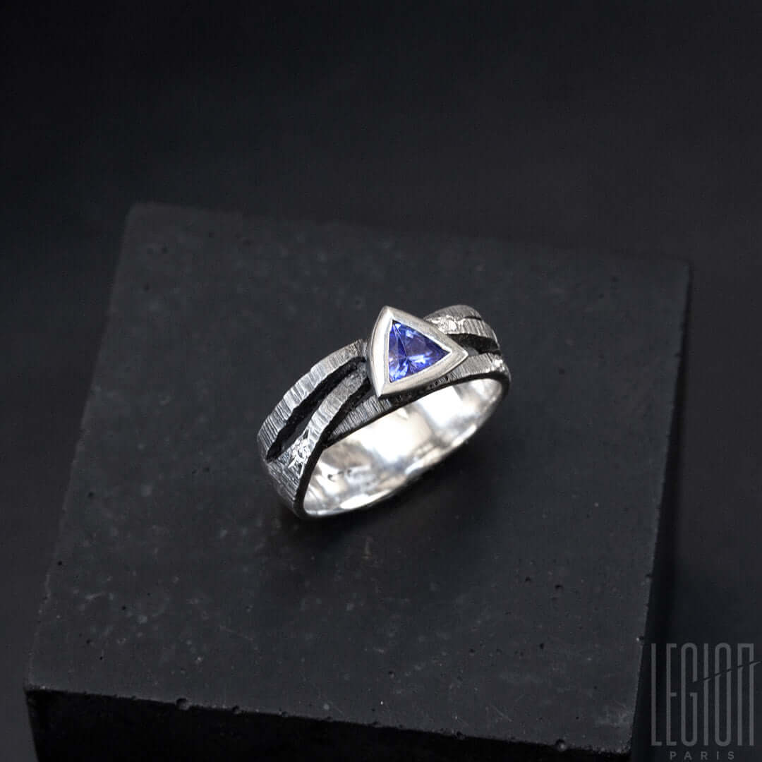 une bague en argent noir texturé composée de 3 brindilles se croisant sur le dessus et d'une tanzanite bleue de forme triangulaire sertie en clos avec sur le corps de bagues des diamants gris parsemés