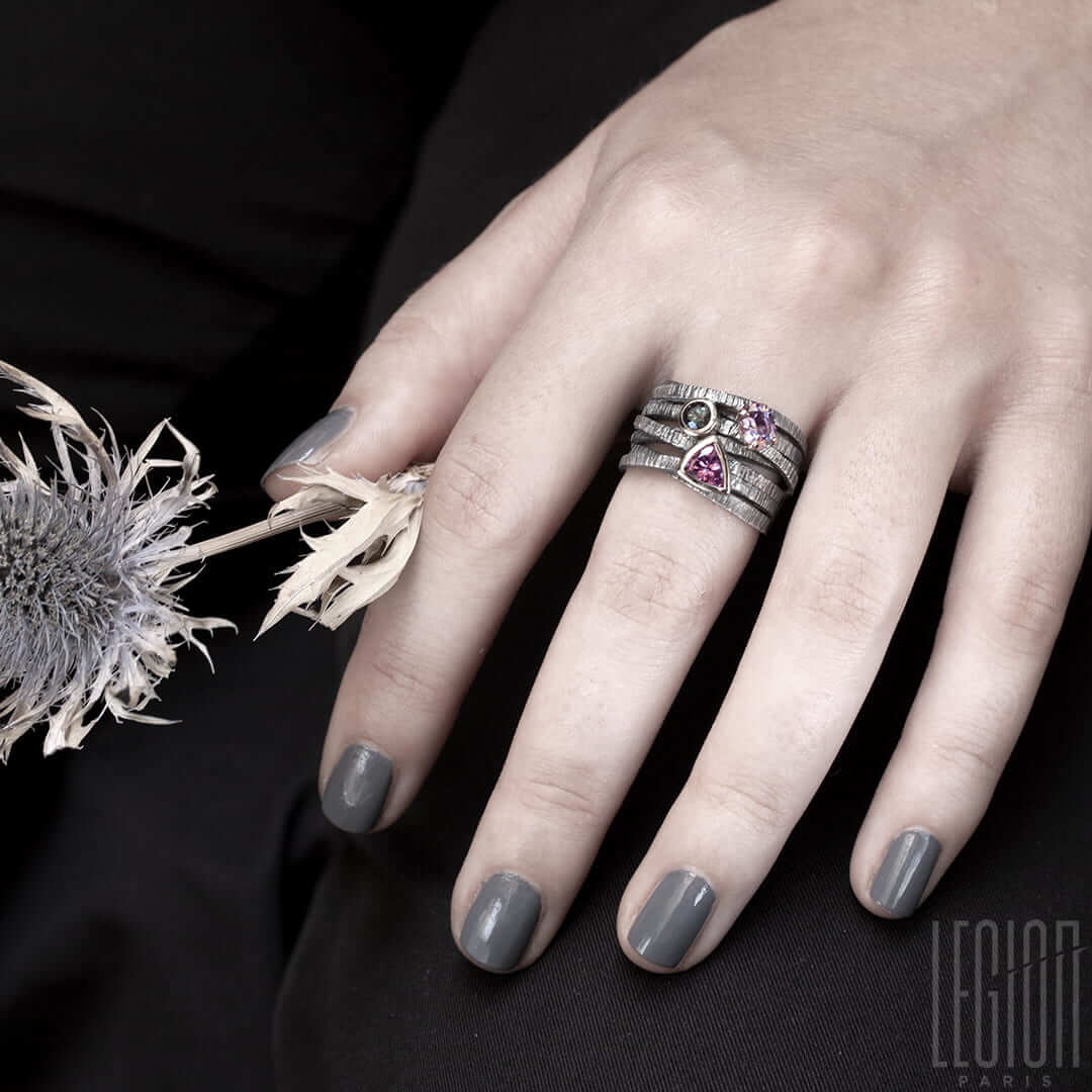 Main de femme portant une Chevalière large avec des brindilles d'argent noir texturées et entrecroisées et 3 pierres sur le dessus. Un spinelle triangle rose, une tourmaline bleue ronde , les deux sont serties clos avec de l'or blanc, et une tourmaline mauve ovale sertie en griffes dans un châton en or rouge