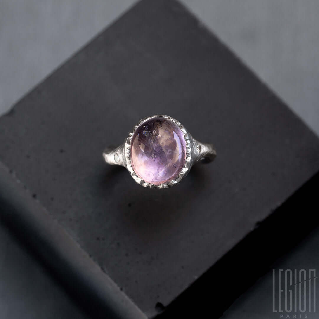 vue d'en haut d'une bague en or blanc avec une pierre mauve ovale et deux diamants gris