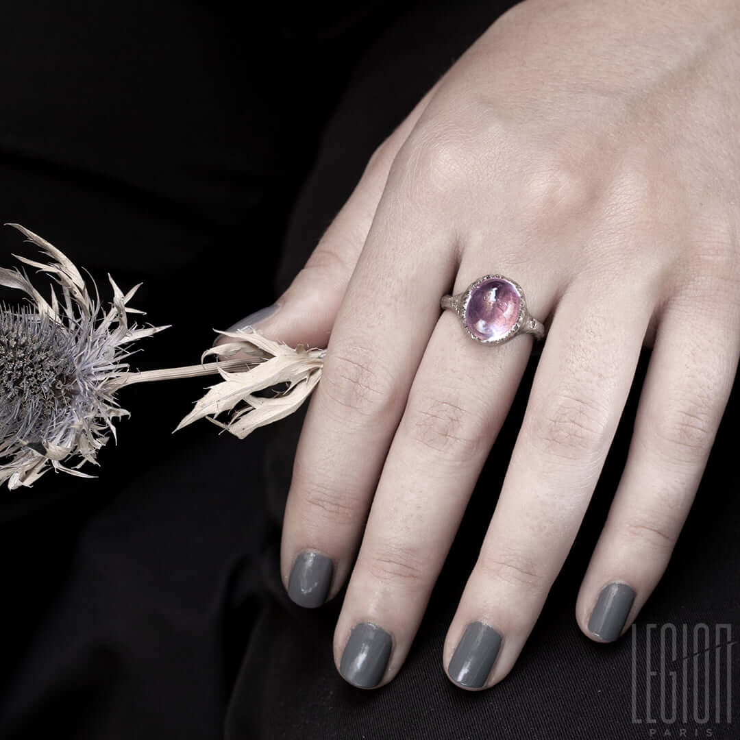 Main de femme portant une Bague chevalière en or blanc texturé avec un gros cabochon de tourmaline rose serti avec des petites griffes qui ressemblent à une coquille d'oeuf