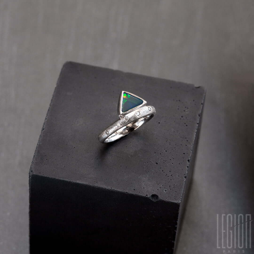 Bague en or blanc texturé avec des diamants gris placés aléatoirement et une opale noire triangle avec des feux vert et orange