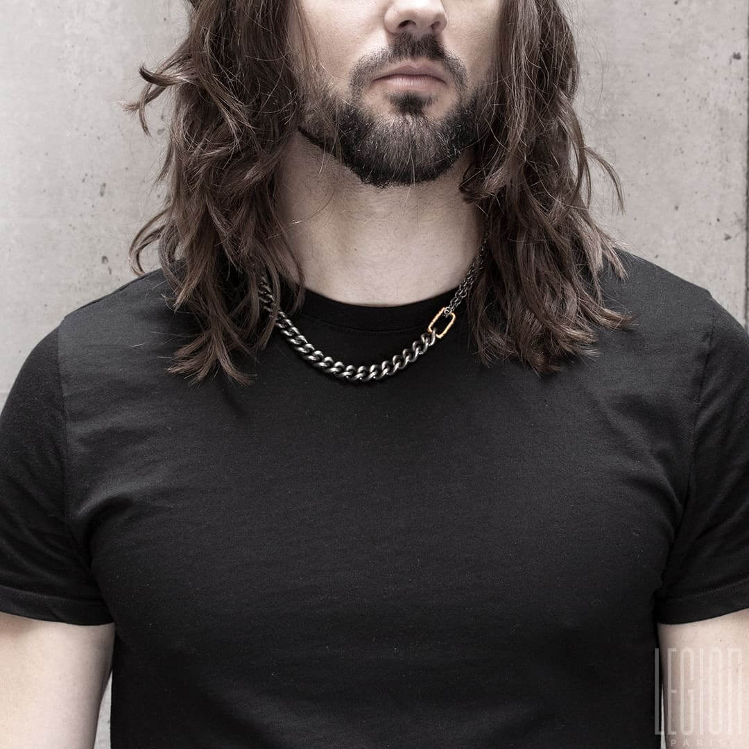 homme aux cheveux longs portant un tee shirt noir et un collier en argent noir et or rouge avec une grosse chaine gourmette