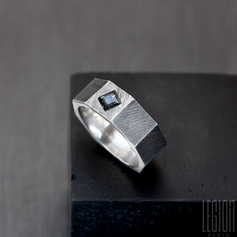 Vue de côté d'un anneau en forme d'ecrou en argent noir avec une pierre gris bleue carrée. L'anneau est strié. 