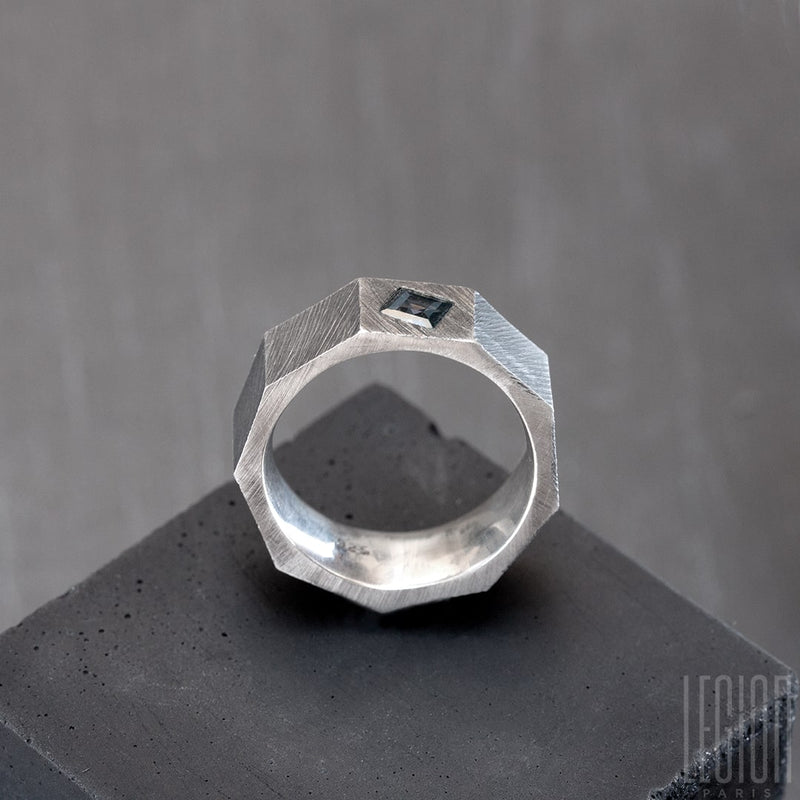 Vue de côté d'un anneau en forme d'ecrou en argent noir avec une pierre gris bleue carrée. L'anneau est strié. 