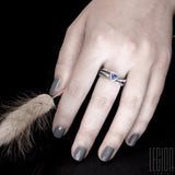 main de femme aux ongles gris portant une bague en argent noir texturé composée de 3 brindilles se croisant sur le dessus et d'une tanzanite bleue de forme triangulaire sertie en clos. 