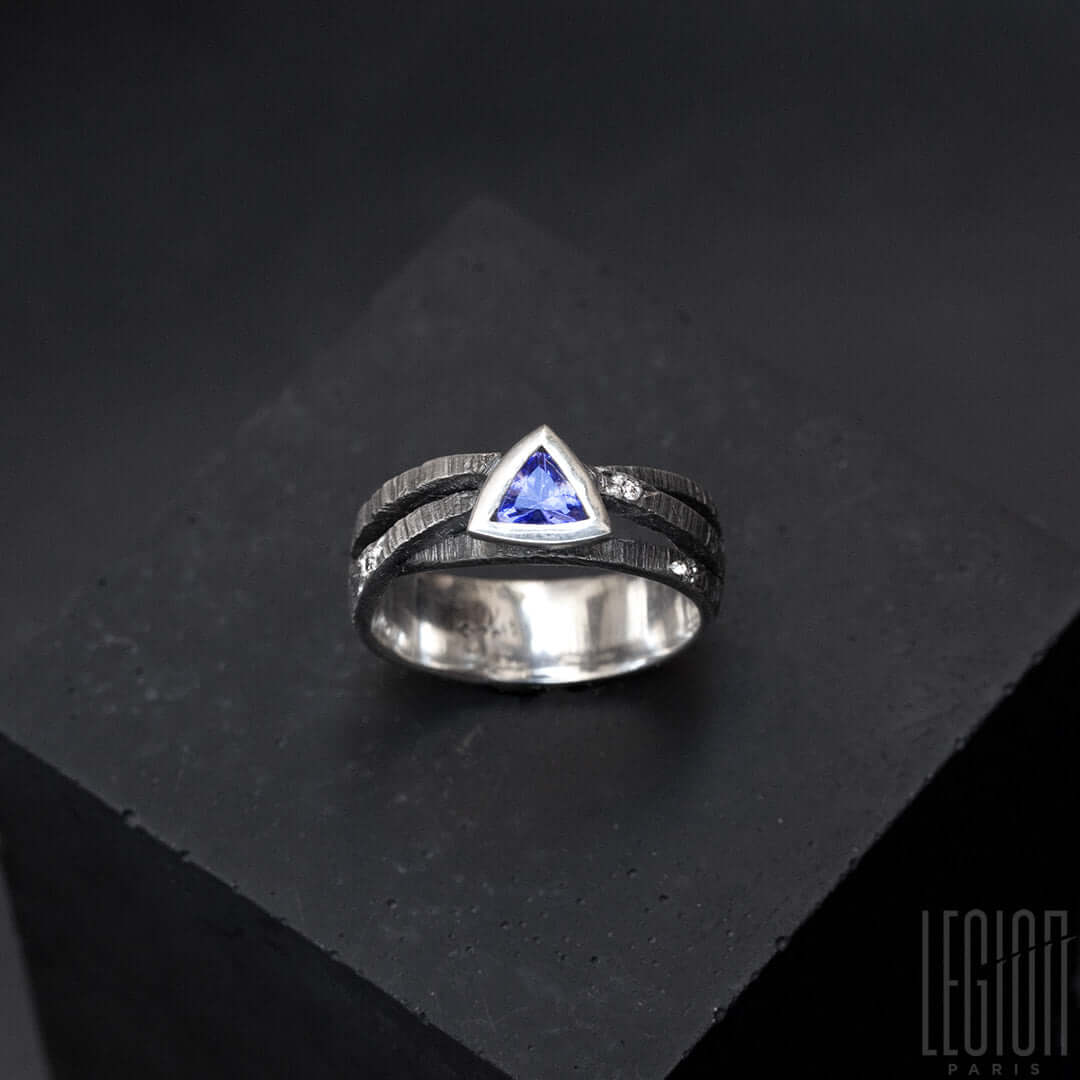 une bague en argent noir texturé composée de 3 brindilles se croisant sur le dessus et d'une tanzanite bleue de forme triangulaire sertie en clos avec sur le corps de bagues des diamants gris parsemés 