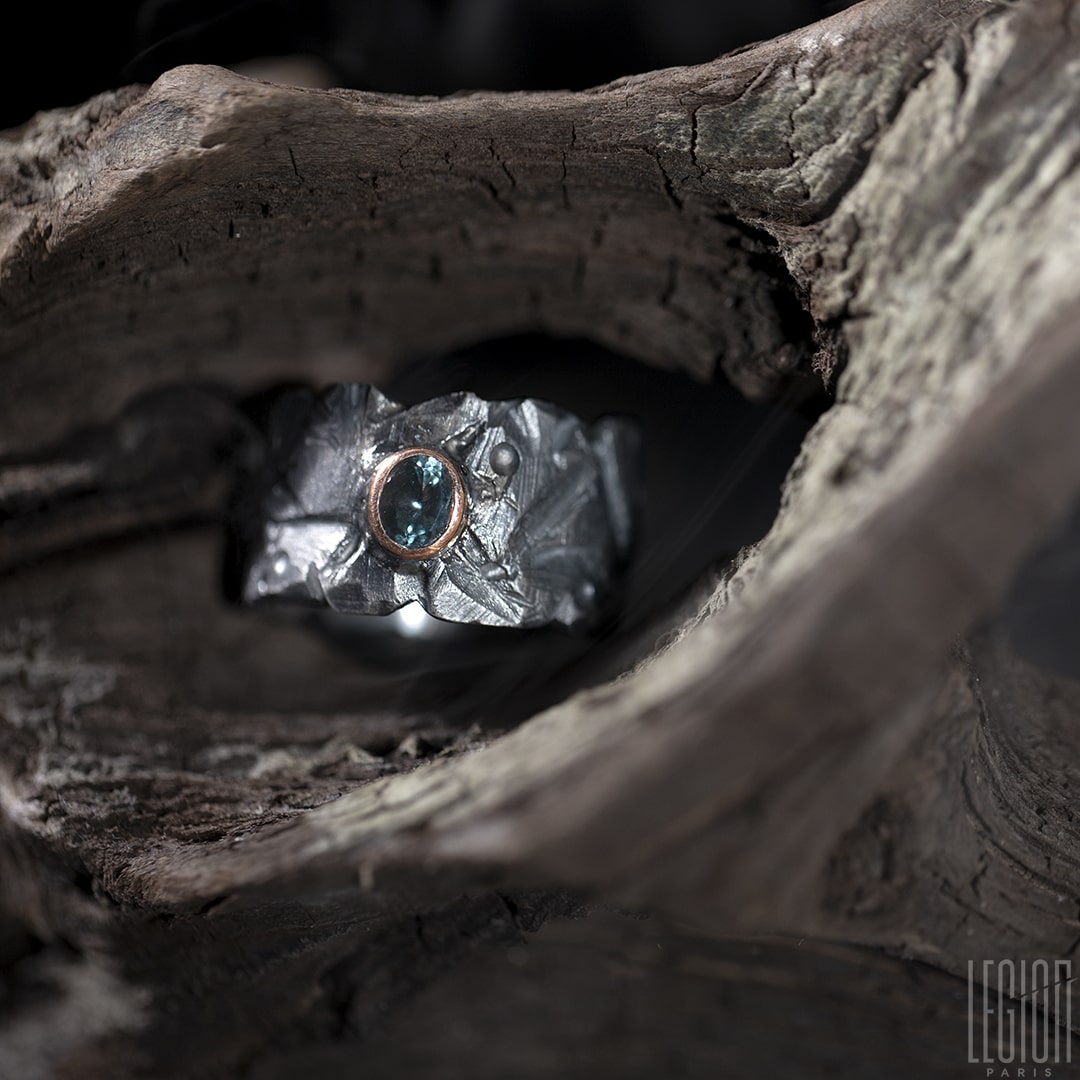 bague large et texturée en argent noir posée sur une racine. Une pierre ovale bleue grise est sertie en clos sur la bague dans de l'or rouge. 