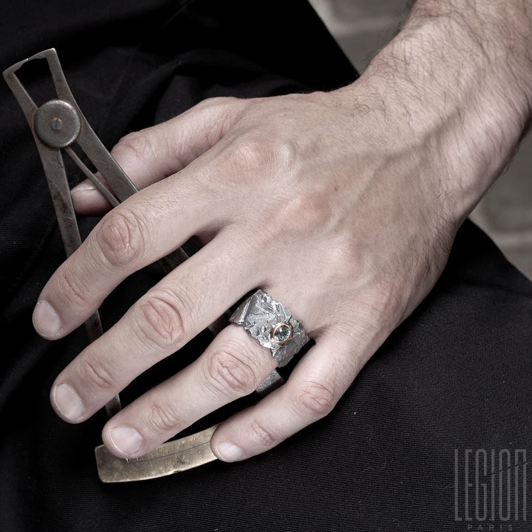 Main d'homme portant une bague très large en argent noir texturé avec une pierre ovale, un spinelle gris vert, serti en clos en or rouge. 