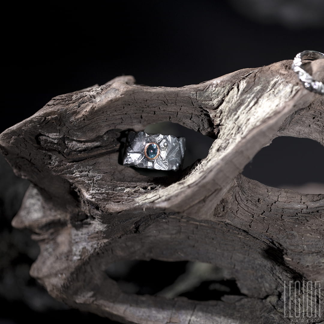 bague large et texturée en argent noir posée sur une racine. Une pierre ovale bleue grise est sertie en clos sur la bague dans de l'or rouge. 
