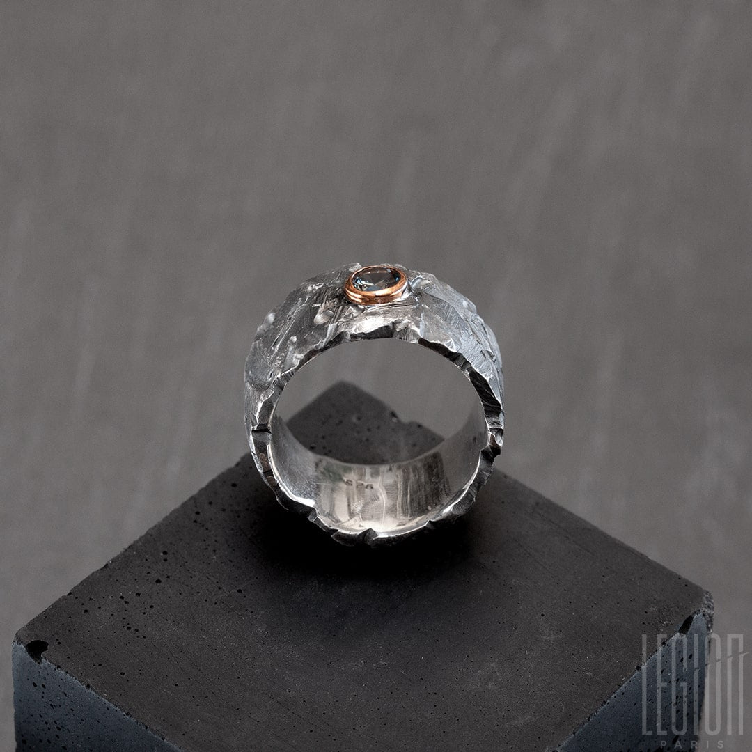 Vue de côté d'unen bague très large en argent noir texturé avec une pierre ovale, un spinelle gris vert, serti en clos en or rouge. 