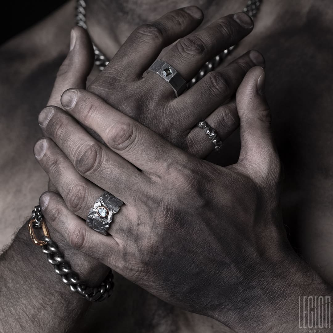 mains d'homme portant un assortiment de bijoux en argent noir avec des chaines gourmettes, des bagues avec des pierres