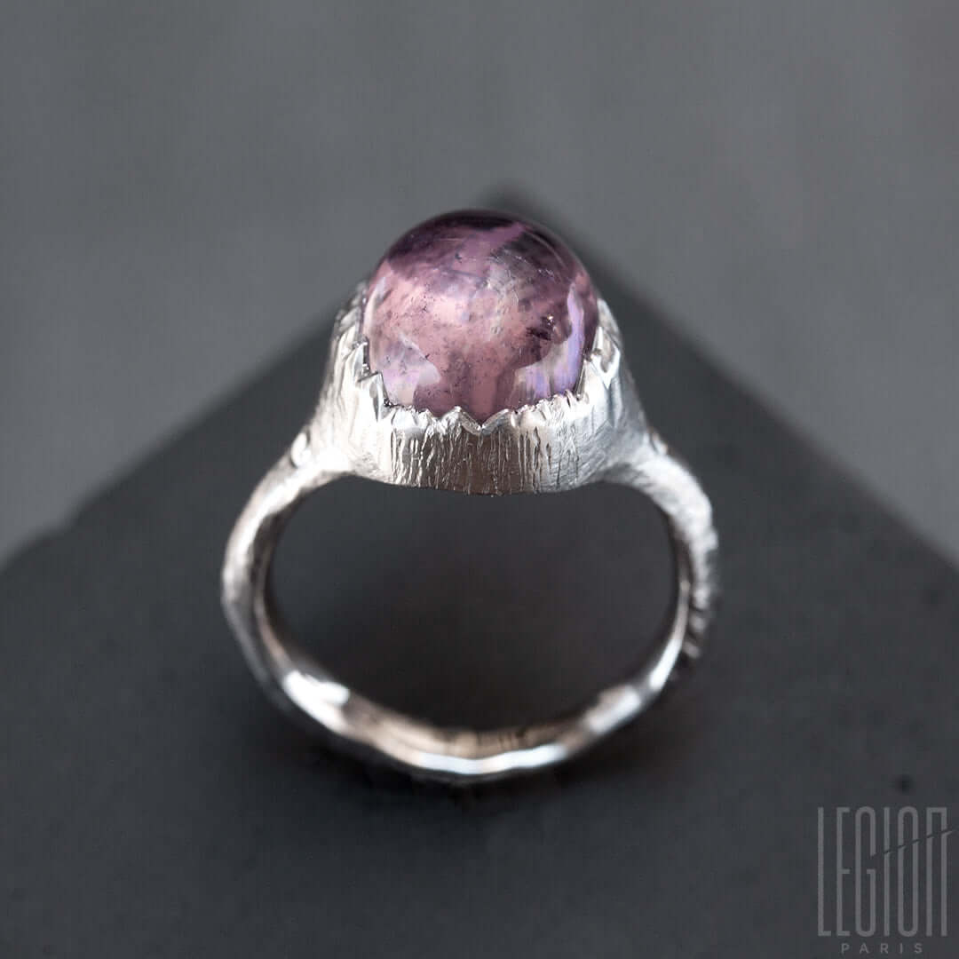 vue de côté d'une bague massive en or blanc avec un gros cabochon de tourmaline rose mauve serti en clos mais avec un effet dechiré