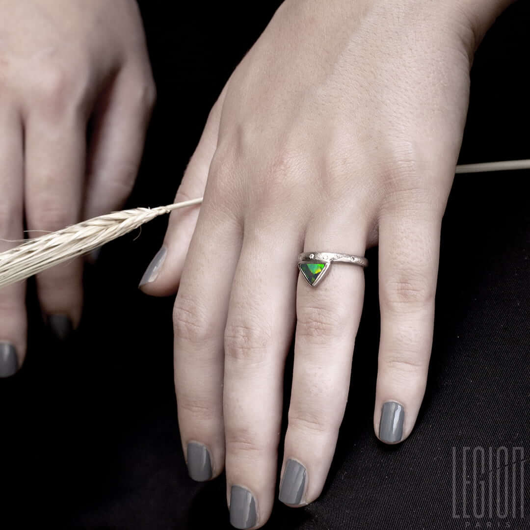 main de femme portant une bague en or blanc texturé avec des diamants gris placés aléatoirement et une opale noire triangle avec des feux vert et orange