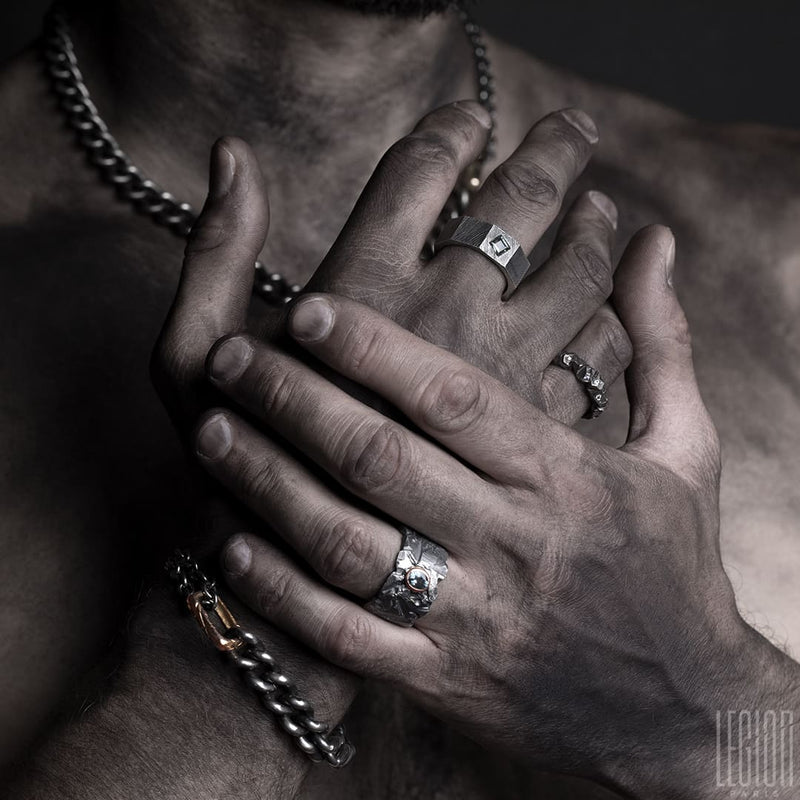 mains d'homme portant un assortiment de bijoux en argent noir avec des chaines gourmettes, des bagues avec des pierres